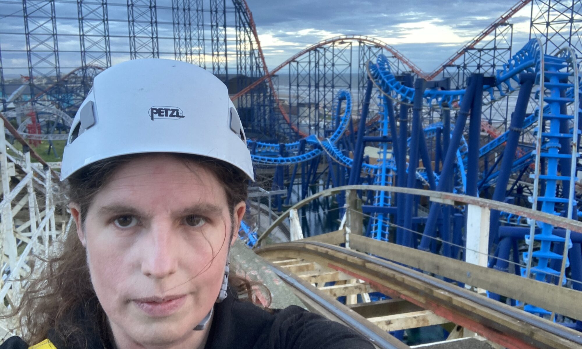 Photo of Katy wearing safety hat and harness, standing on top of the Big Dipper rollercoaster in Blackpool with view of Big One, Infusion and Big Dipper in background